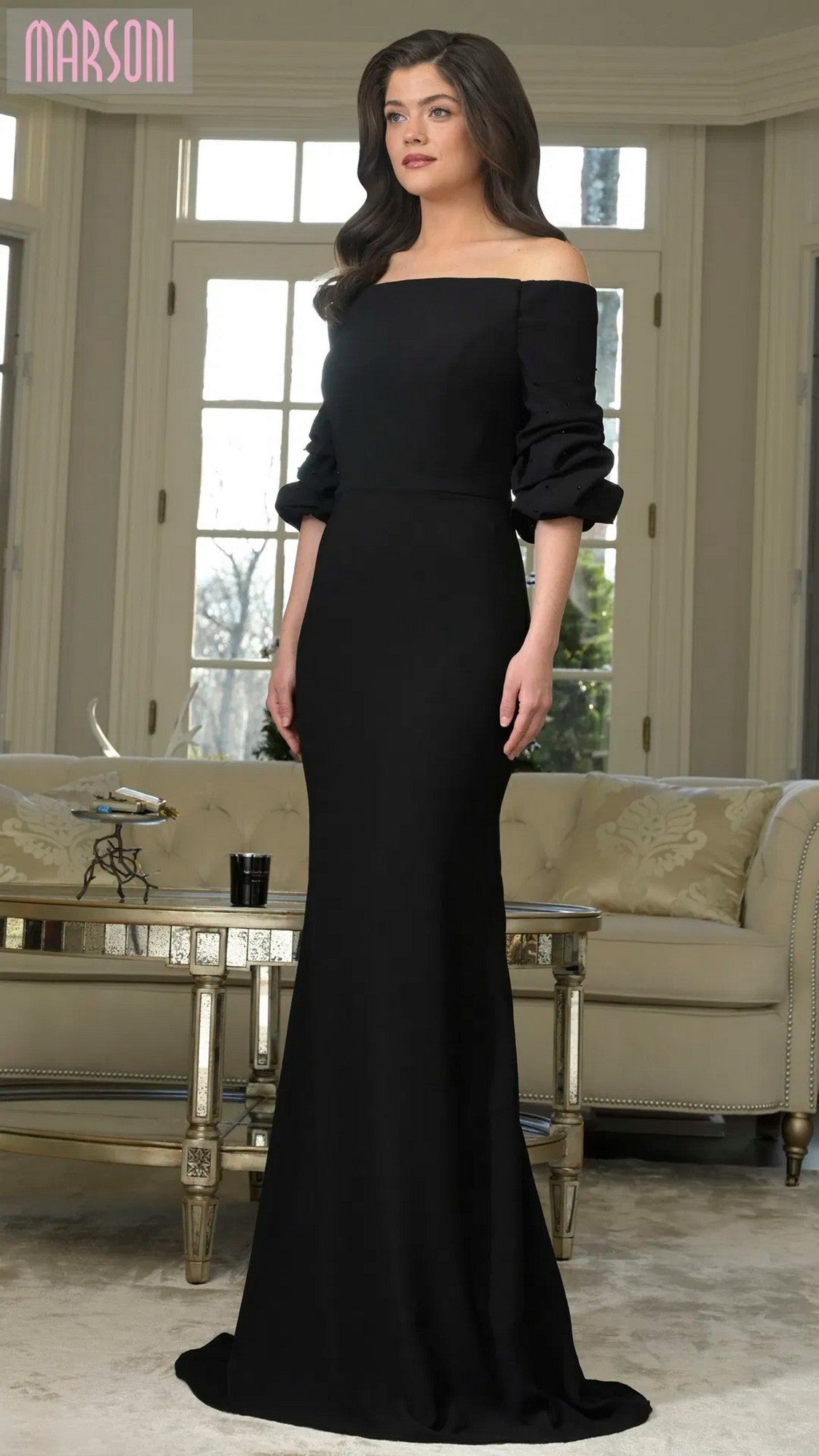 A woman in a black Marsoni MV1337 long formal dress with off-the-shoulder ruffled sleeves stands in a living room with large windows, a beige sofa, and a glass coffee table.